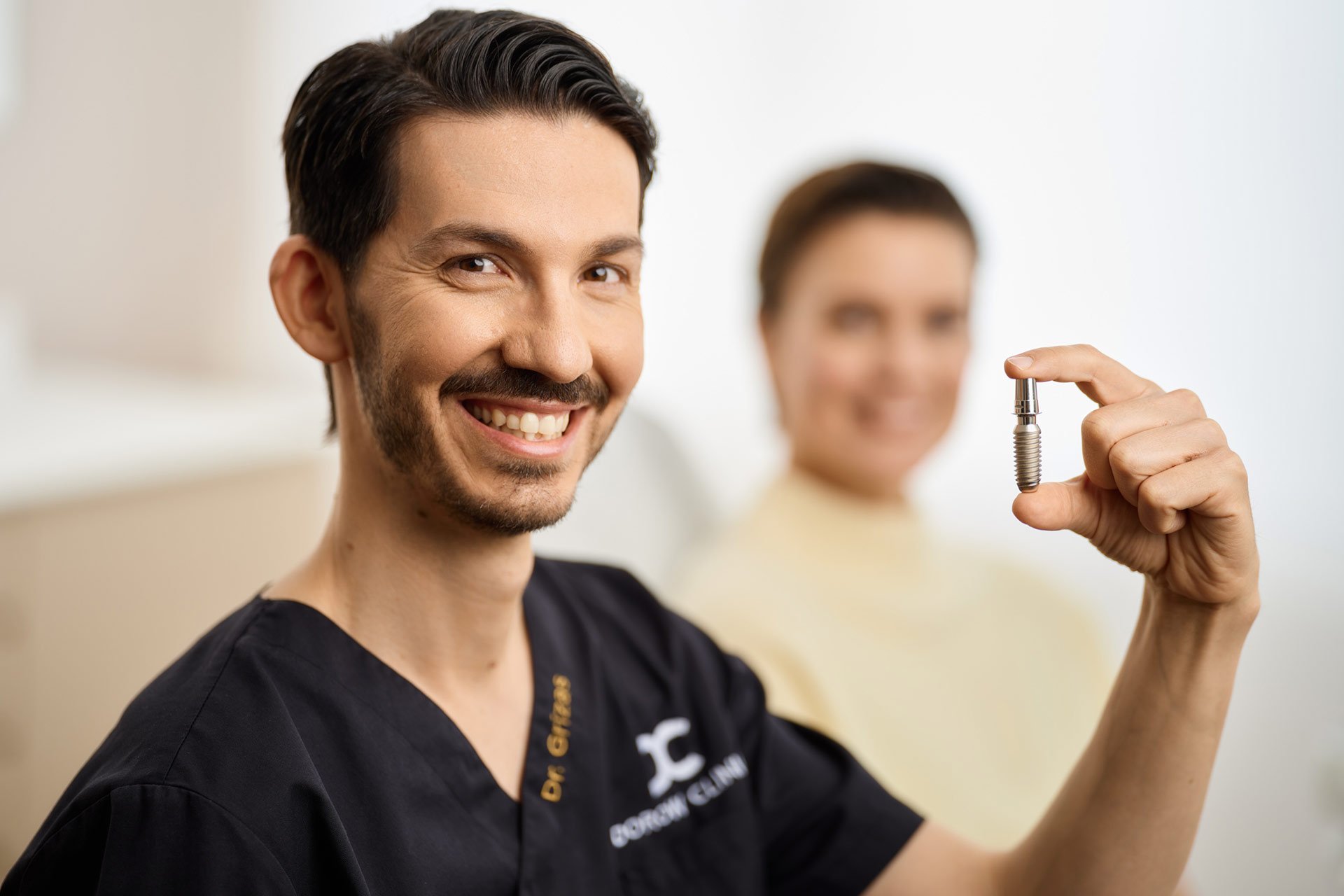 Dr Eleftherios Grizas von der Dorow Clinic mit einem Zahnimplantat / Zahnersatz in den Händen. Im Hintergrund sitzt eine Zahnpatientin auf dem Zahnarztstuhl